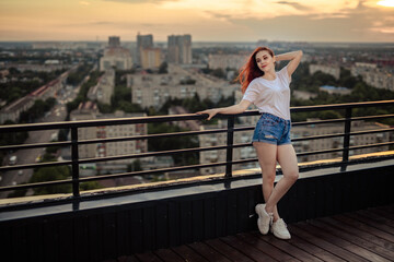 Portrait of a red-haired girl with long hair on the roof of a high house at sunset overlooking the city.