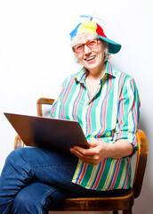 Portrait of mature smiling woman in multicolored cap with propeller and glasses sitting at laptop on a chair on white background