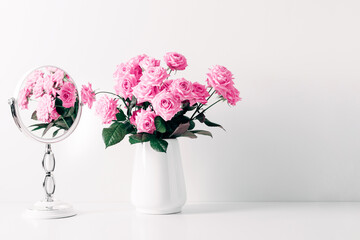 Pink roses in vase reflected in mirror. Light white bathroom decor, mirror, pink roses flowers on white shelf. Elegant decor bathroom interior.