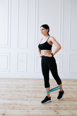 Obraz premium The girl athlete in black tight-fitting clothes put her hands on her waist and does exercises with an elastic expander on her ankles.