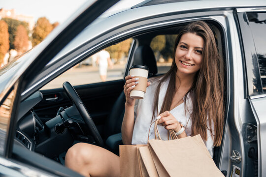 Beautiful young happy smiling woman with packages after shopping drink coffee sitting in her car . Coffee break concept.
