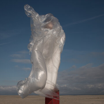 Girl in white dress like a  modern classic statue wrapped in plastic on the beach under a blue sky