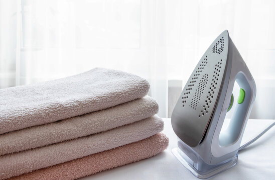 An electric iron and a stack of ironed towels on a light surface. Laundry ironing, economics, household. Horizontal orientation, selective focus.