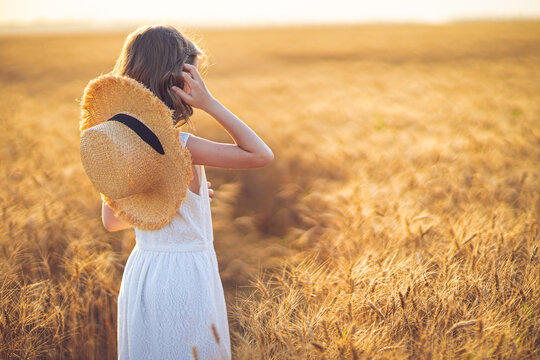 Fashion photo of a little girl in white dress and straw hat at the evening wheat field. Back view
