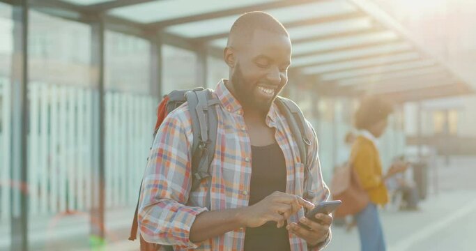 Young handsome African American man with backpack texting message on smartphone and smiling cheerfully at train station. Attractive male traveller tapping and scrolling on mobile phone at bus stop.