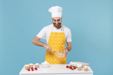 Smiling young male chef or cook baker man in apron white t-shirt toque chefs hat cooking at table isolated on blue background. Cooking food concept. Mock up copy space. Whipping beating eggs in bowl.