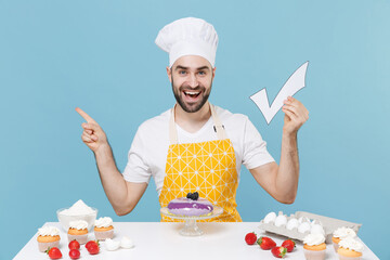 Excited young bearded male chef or cook baker man in apron white t-shirt toque chefs hat cooking at table isolated on blue background. Cooking food concept Hold check mark point index finger aside up.