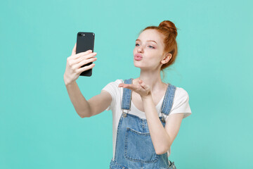 Pretty young readhead girl in casual denim clothes posing isolated on blue turquoise background. People lifestyle concept. Mock up copy space. Doing selfie shot on mobile phone, blowing send air kiss.