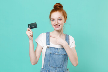 Smiling young readhead girl in casual denim clothes isolated on blue turquoise wall background studio portrait. People lifestyle concept. Mock up copy space. Pointing index finger on credit bank card.