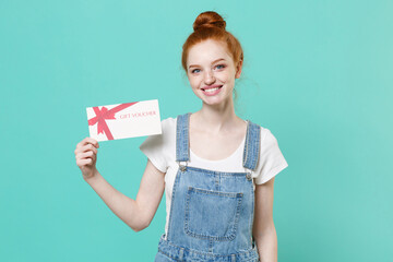 Smiling young readhead girl in casual denim clothes isolated on blue turquoise wall background studio portrait. People sincere emotions lifestyle concept. Mock up copy space. Hold gift certificate.