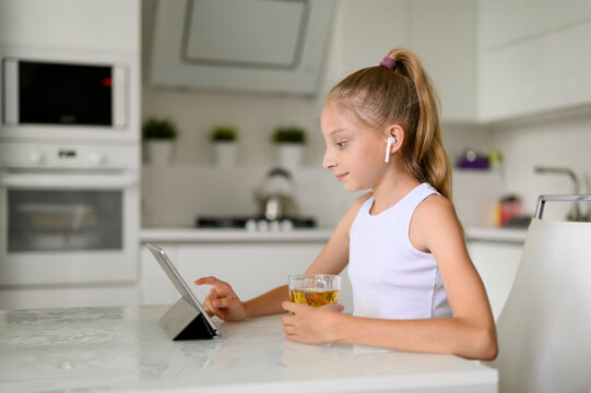 Girl sitting in the kitchen at the table with a glass of lemonade. Girl with wireless headphones communicates on the Internet using a tablet.