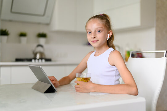 A girl in a white T-shirt and long blond hair sits at home at the table. Nearby is a tablet. A girl holds a glass of juice in her hand. Girl looking at the camera and smiling.