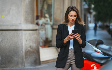elegant girl in business suit standing at parking mopeds on street european city and using mobile...