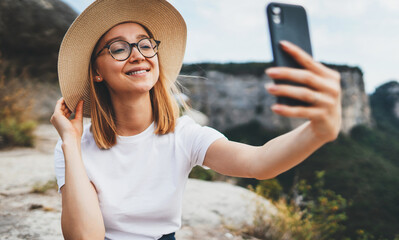 smiling girl traveler in hat communicates by video call on mobile phone ralax on top of mountain landscape, female hipster tourist takes photo selfie on cellphone on background of nature outside