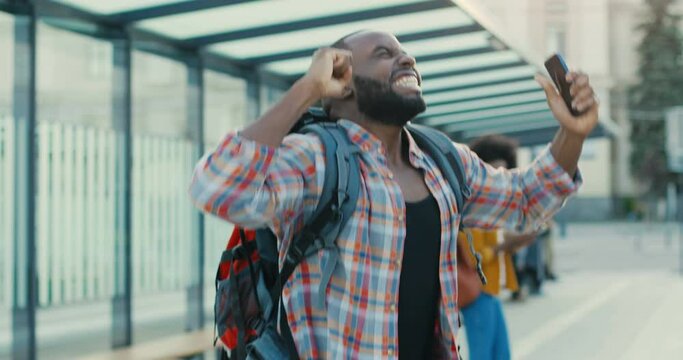 Young African American Cheerful Handsome Man Traveller With Backpack Tapping And Scrolling On Smartphone At Bus Stop. Happy Guy Reading Message Or News On Mobile Phone And Jumping From Happiness.