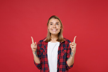 Smiling young blonde woman girl in casual checkered shirt posing isolated on red background studio portrait. People sincere emotions lifestyle concept. Mock up copy space. Pointing index fingers up.