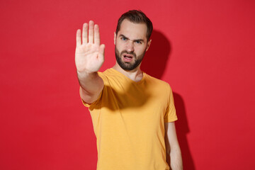 Naklejka premium Displeased young bearded man guy in casual yellow t-shirt posing isolated on red wall background studio portrait. People emotions lifestyle concept. Mock up copy space. Showing stop gesture with palm.
