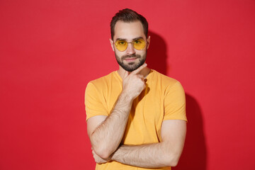 Obraz premium Smiling young bearded man guy in casual yellow t-shirt glasses posing isolated on red wall background studio portrait. People emotions lifestyle concept. Mock up copy space. Put hand prop up on chin.