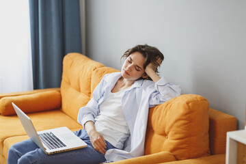 Tired young girl in casual clothes sit on couch spending time in living room at home. Rest relax good mood leisure lifestyle concept. Work on laptop pc computer, put hand on head, keeping eyes closed.