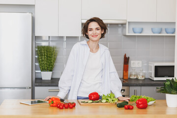 Smiling charming young brunette housewife woman girl in white casual clothes preparing vegetable salad cooking food in light kitchen at home. Dieting healthy lifestyle concept. Mock up copy space.