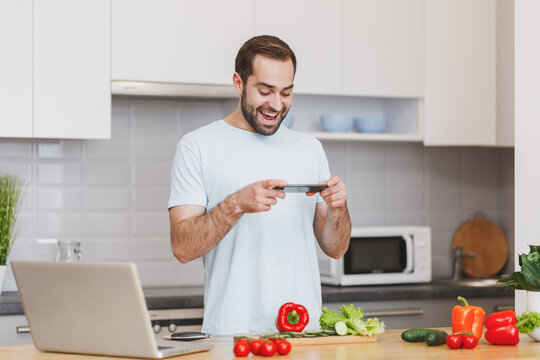 Funny young bearded man guy in white t-shirt taking pictures on mobile phone preparing vegetable salad cooking food in light kitchen at home. Dieting healthy lifestyle concept. Mock up copy space.