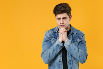 Pensive young man guy 20s in casual denim jacket posing isolated on yellow background studio portrait. People sincere emotions lifestyle concept. Mock up copy space. Holding hands folded in prayer.