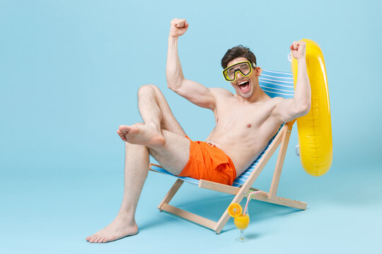 Happy young man guy in orange shorts swimming goggles sit on deck chair isolated on blue background studio. People summer vacation rest lifestyle concept. Mock up copy space. Doing winner gesture. - Powered by Adobe