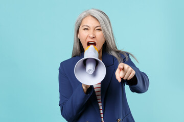 Angry gray-haired business woman in blue suit posing isolated on blue background. Achievement career wealth business concept. Mock up copy space. Scream in megaphone, pointing index finger on camera.