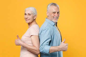 Side view of smiling elderly gray-haired couple woman man in casual clothes posing isolated on yellow background. People lifestyle concept. Mock up copy space. Standing back to back showing thumbs up.