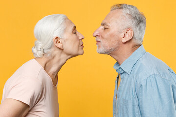 Side view of pretty elderly gray-haired couple woman man in casual clothes posing isolated on yellow wall background studio. People lifestyle concept. Mock up copy space. Kissing, keeping eyes closed.