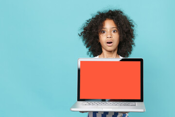 Fototapeta na wymiar Shocked little african american kid girl 12-13 years old in striped clothes isolated on blue background. Childhood lifestyle concept. Mock up copy space. Hold laptop computer with blank empty screen.