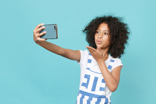 Pretty little african american kid girl 12-13 years old in striped clothes isolated on blue background studio. Childhood lifestyle concept. Doing selfie shot on mobile phone, blowing sending air kiss.