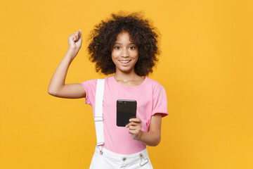 Joyful little african american kid girl 12-13 years old in pink t-shirt isolated on yellow background studio. Childhood lifestyleconcept. Mock up copy space. Using mobile phone doing winner gesture.