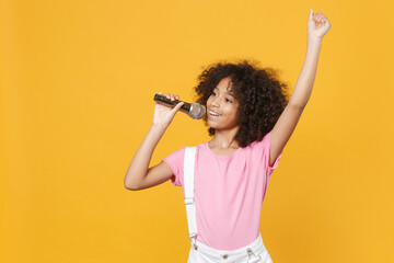 Pretty little african american kid girl 12-13 years old in pink t-shirt isolated on yellow wall background studio. Childhood lifestyle concept. Mock up copy space. Sing song in microphone rising hand.