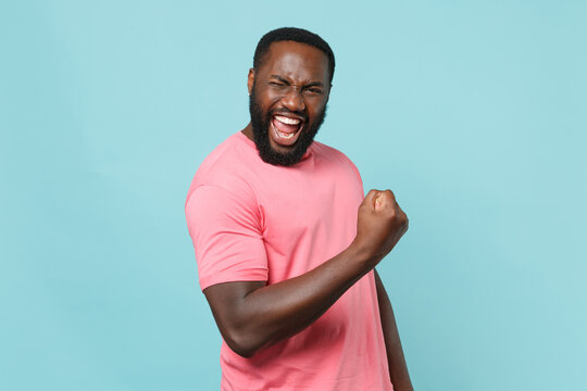 Happy young african american man guy in casual pink t-shirt posing isolated on pastel blue background studio portrait. People emotions lifestyle concept. Mock up copy space. Doing winner gesture.