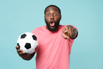 Shocked african american man football fan in pink t-shirt isolated on blue background. Sport family leisure concept. Cheer up support favorite team with soccer ball, pointing index finger on camera.