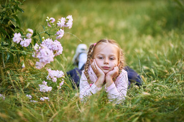 The little girl lies on the green grass near the bush with pink flowers and makes different facial expressions. girl in pink blouse and blond hair