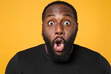 Close up of shocked african american man football fan in casual black t-shirt isolated on yellow background studio portrait. People emotions lifestyle concept. Mock up copy space. Keeping mouth open.