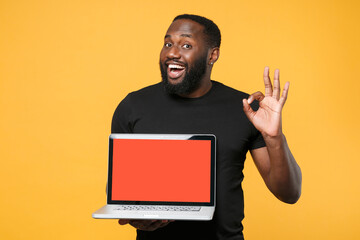 Excited african american man in casual black t-shirt isolated on yellow background. People lifestyle concept. Mock up copy space. Hold laptop pc computer with blank empty screen showing OK gesture.