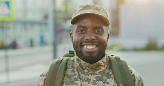 Portrait shot of joyful handsome African American young male soldier in cap with backpack smiling to camera at street. Close up of man militarian with smile outdoor. Military uniform. Going to army.