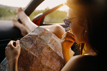 Close-up portrait of a young woman sitting in car in sunset light, holding a map, planning new...