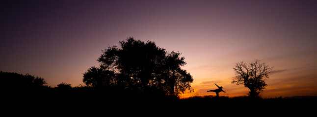 silhouette of tow trees with a girl in the middle making shapes with her body during a sunset with orange color clouds. 