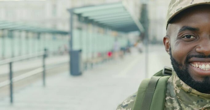 Portrait of joyful African American handsome young man soldier in cap with backpack smiling at street. Close up of half face of male militarian with happy smile outdoor at bus stop. Military uniform.