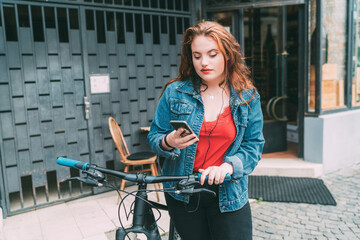 Fototapeta na wymiar Portrait of red curled long hair caucasian teen girl on the city street walking with bicycle using the smartphone with earphones. Natural people beauty urban life concept image.