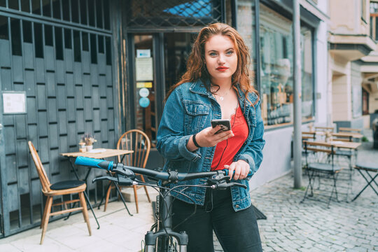 Red curled long hair caucasian teen girl on the city street walking with bicycle using the smartphone with earphones fashion portrait. Natural people beauty urban life concept image.