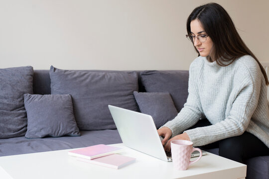 Young Brunette Woman Working On A Laptop On A Grey Couch And White Background