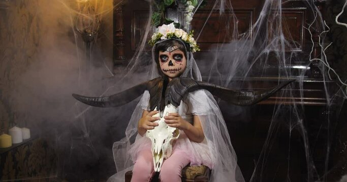 Portrait of girl dressed in Halloween Santa muerte costume  holding a goat scull against background of old dark Gothic interior covered with cobwebs and mysterious fog. 4K slow motion 50 fps