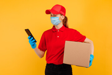 young female courier in a red Cap and t-shirt, with a medical protective mask on her face and gloves. A delivery girl is holding a cardboard box and using a mobile phone. Online order, coronavirus