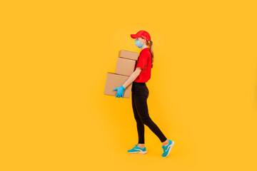 Fototapeta na wymiar Full length, young female courier dressed in a Red cap and t-shirt, uniform, medical protective mask and gloves, holds cardboard boxes and shows a gesture of strength against a yellow background
