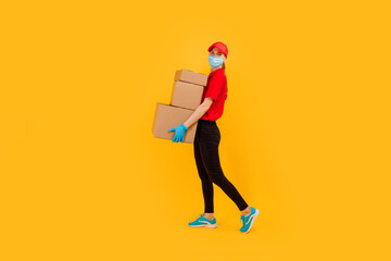 Fototapeta na wymiar Full length, young female courier dressed in a Red cap and t-shirt, uniform, medical protective mask and gloves, holds cardboard boxes and shows a gesture of strength against a yellow background